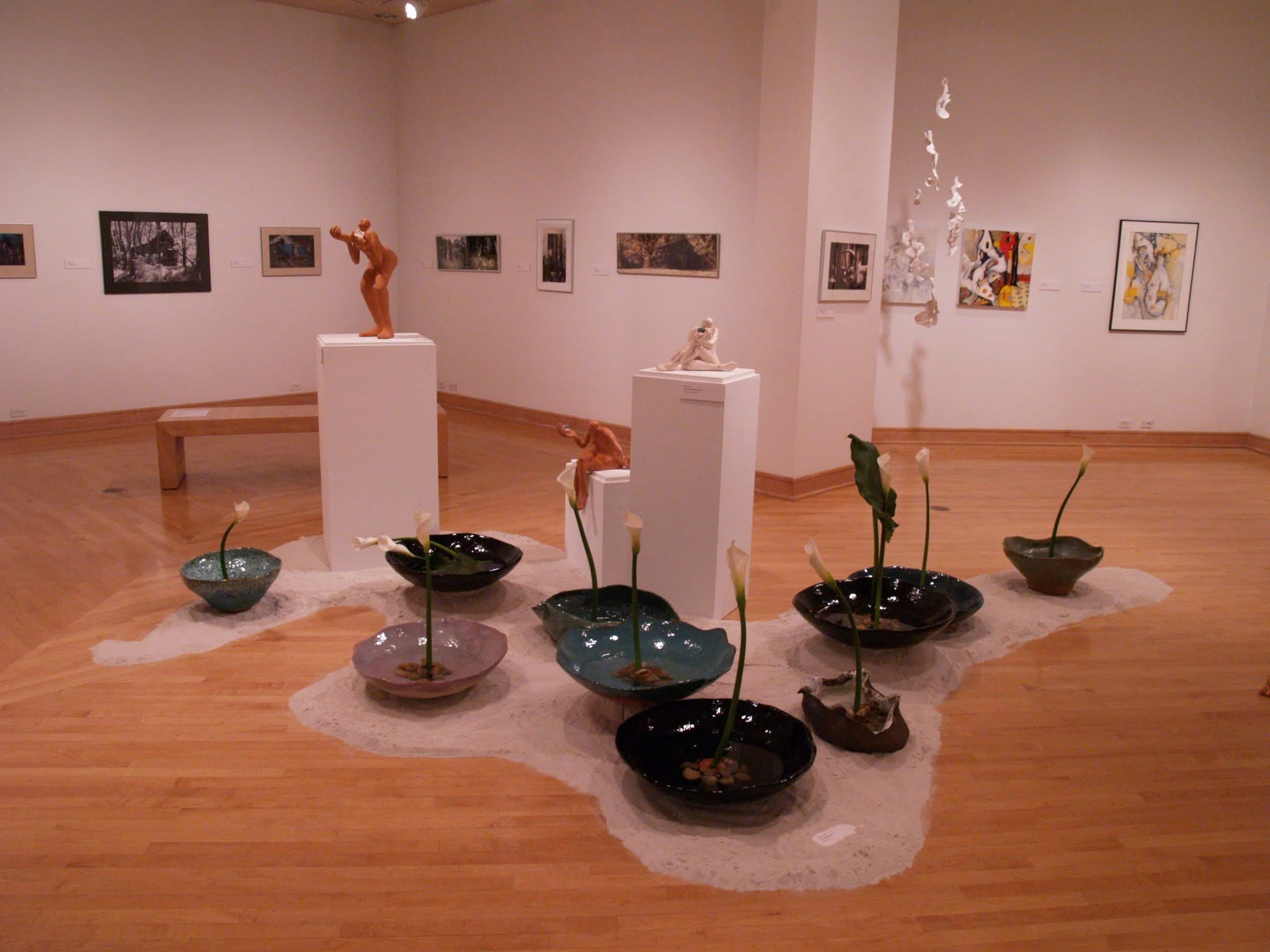 Gallery view of the bowl installation in white sand, with sculpted figures on white plinths behind