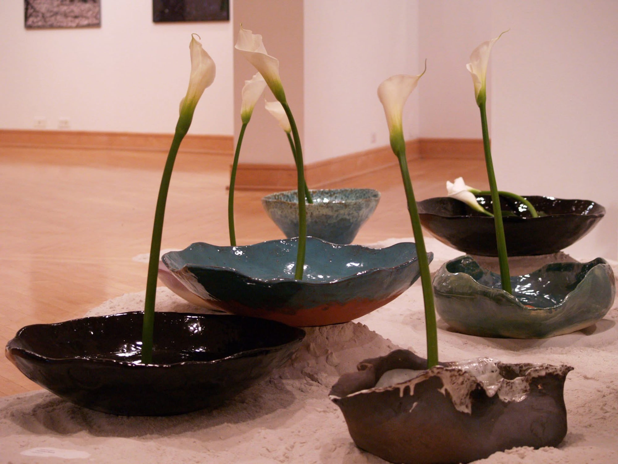 Hand-built bowls in black and teal glaze resting in white sand, each with a tall calla lily