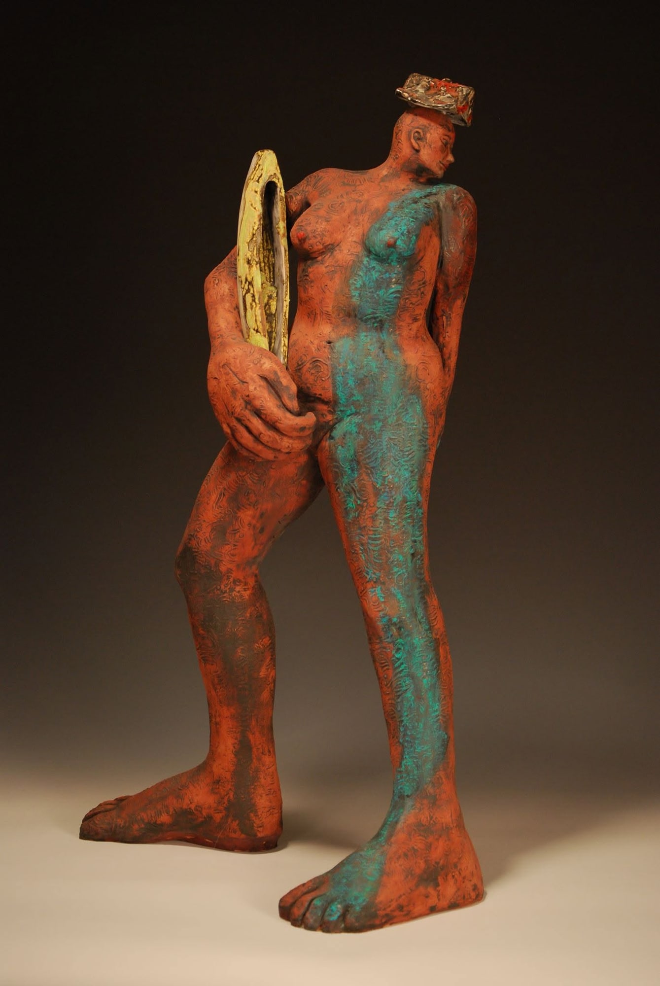 A tall orange-red stoneware figure with a streak of teal glaze down one side, holding a yellow oval like a hand mirror against her body