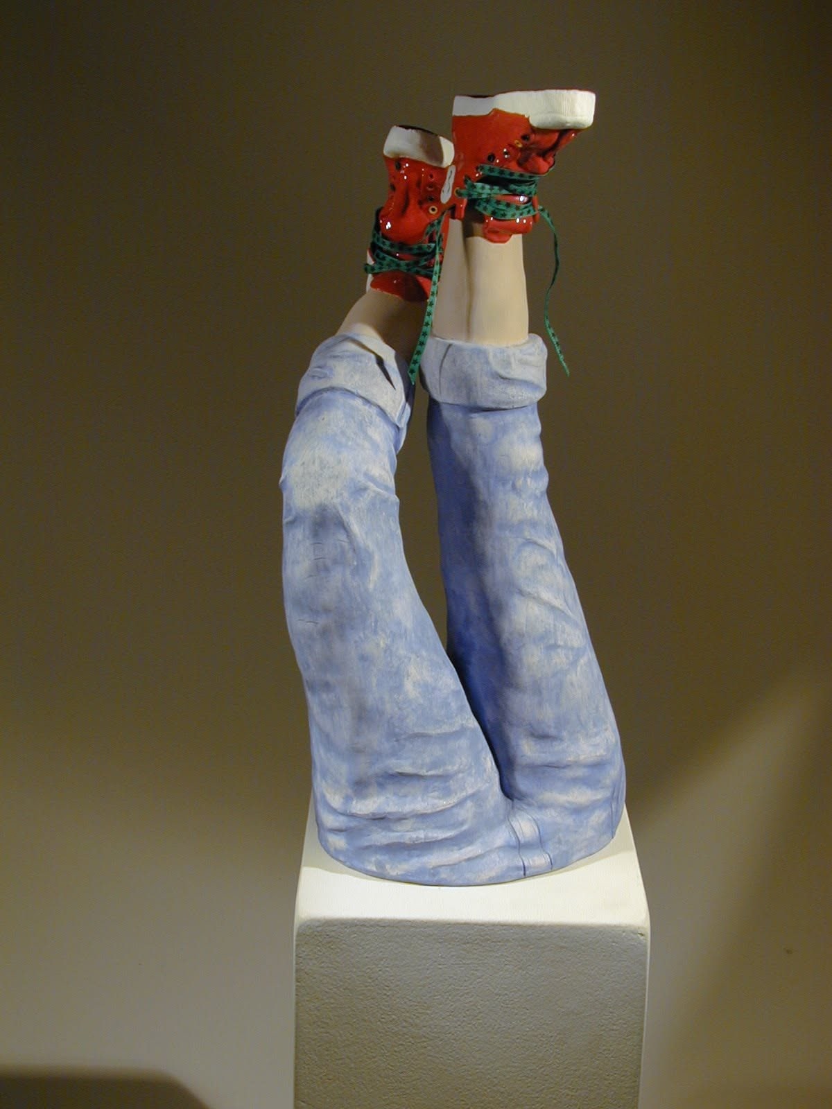Upturned ceramic legs in faded blue jeans, feet in the air in red high-top sneakers with green laces, on a white plinth