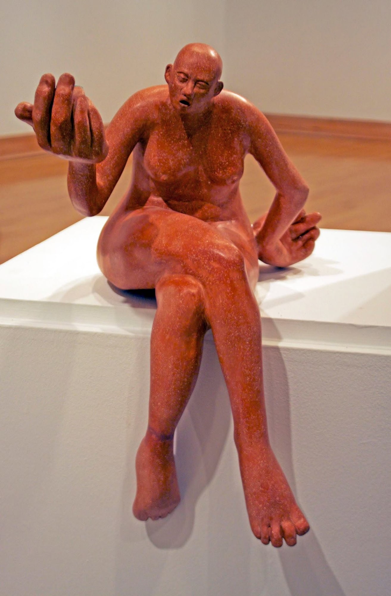A red stoneware figure sitting on the edge of a white plinth, legs dangling, one oversized hand raised and open