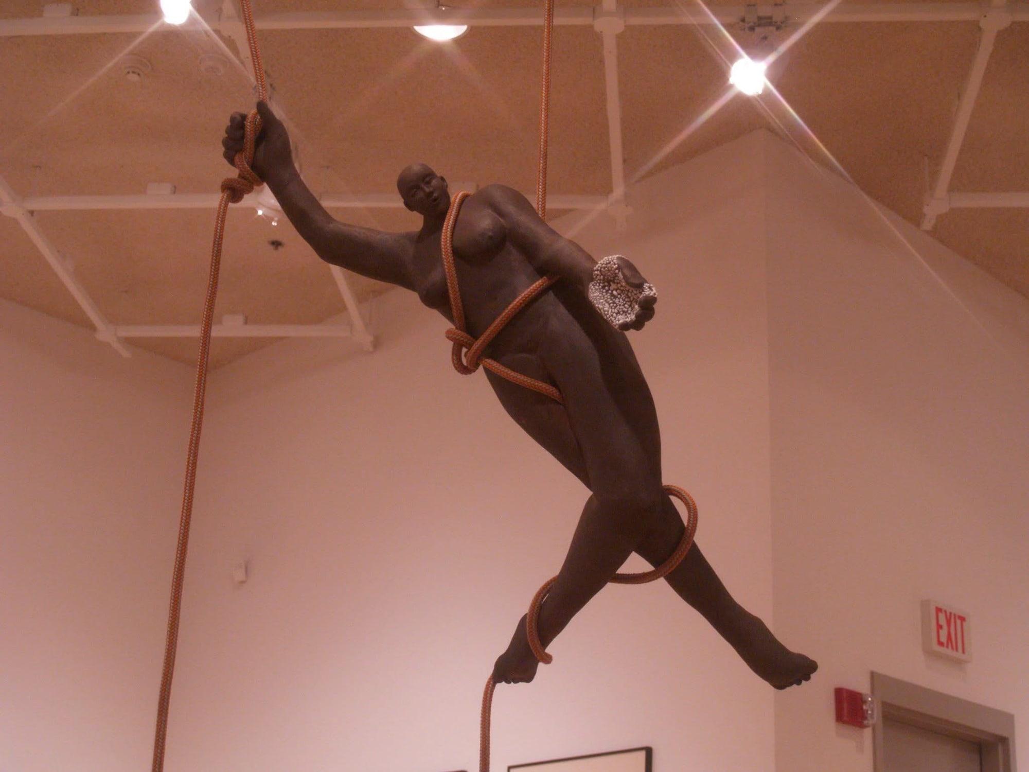 A dark stoneware figure hanging from the gallery ceiling, wrapped and held by orange rope