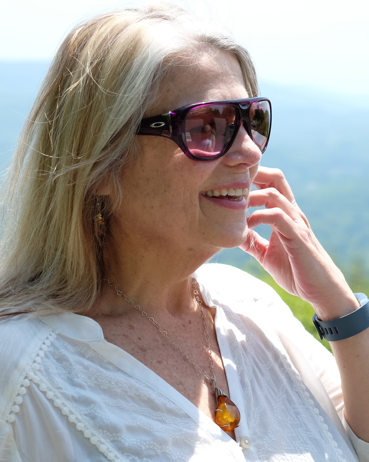 Deborah laughing in purple sunglasses, hand to her cheek, hills behind her